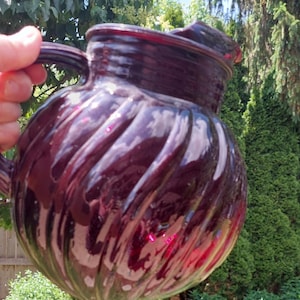 May include: A deep dark red glass pitcher featuring a rounded body with a prominent vertical ribbed or swirled pattern. The pitcher has a wide mouth, a sturdy handle on the side, and a thick rim, reflecting light across its glossy, translucent surface.