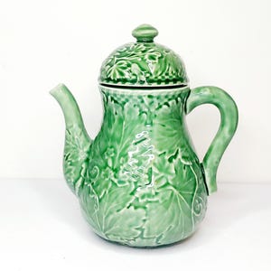 May include: A green ceramic teapot with a lid, spout, and handle. The teapot is decorated with a raised leaf pattern. The lid has a small knob on top. The teapot is a light green color.