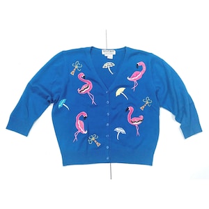 May include: A blue cardigan sweater with a V-neck and button closure. The sweater features embroidered pink flamingos, umbrellas, and palm trees. The sleeves are three-quarter length.