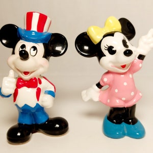 May include: A pair of ceramic figurines of Mickey Mouse and Minnie Mouse. Mickey is wearing a blue and red hat and a blue and red outfit. Minnie is wearing a pink dress with white polka dots and a yellow bow.