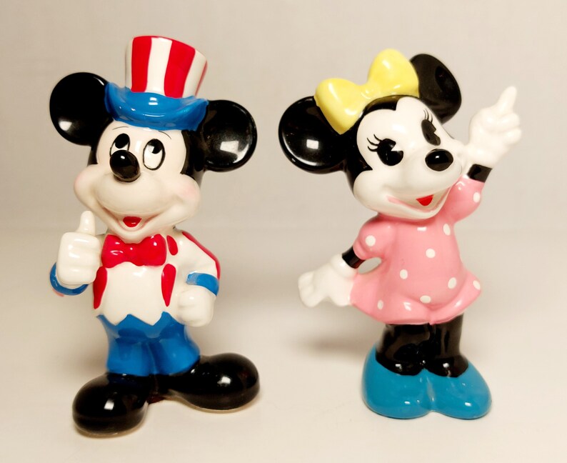 May include: A pair of ceramic figurines of Mickey Mouse and Minnie Mouse. Mickey is wearing a blue and red hat and a blue and red outfit. Minnie is wearing a pink dress with white polka dots and a yellow bow.