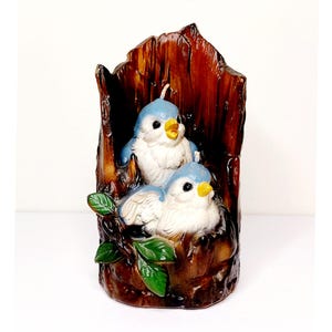 May include: A candle shaped like a tree trunk with two bluebirds perched on a branch. The birds have white bellies and yellow beaks. The tree trunk is brown and has a rough texture.
