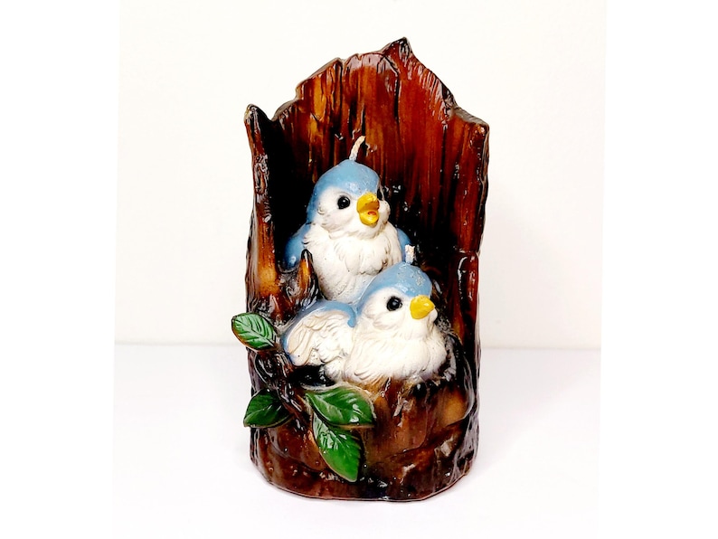 May include: A candle shaped like a tree trunk with two bluebirds perched on a branch. The birds have white bellies and yellow beaks. The tree trunk is brown and has a rough texture.
