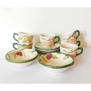 May include: A set of cream-colored ceramic teacups, saucers, and bowls with hand-painted fruit designs. The cups have green handles and the rims of the bowls are green. The fruit includes peaches, cherries, and plums.