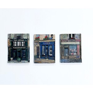 May include: Three rectangular wall plaques depicting Parisian storefronts. The left plaque shows a dark green restaurant labeled Chez Jerome Restaurant 62. The center plaque features a blue bookstore labeled Livres. The right plaque shows a shop labeled France Soir.
