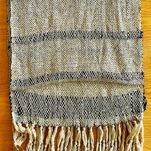 May include: Woven scarf with a cream and black striped pattern. The scarf has a fringed edge and a tag attached. The textile is made of a woven material. The scarf is folded on a wooden surface.