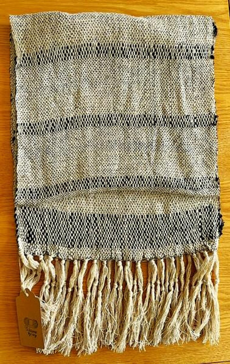 May include: Woven scarf with a cream and black striped pattern. The scarf has a fringed edge and a tag attached. The textile is made of a woven material. The scarf is folded on a wooden surface.