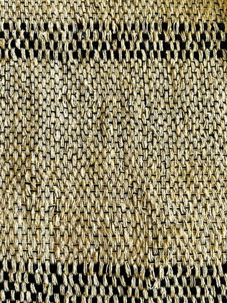 May include: Close-up of a woven textile with a textured pattern. The fabric is primarily beige with horizontal bands of black. The weave creates a grid-like design, showcasing the natural fibers and craftsmanship.