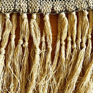 May include: Close-up of a woven textile with a black and beige geometric pattern at the top. Below, long, knotted beige fringe hangs down. The textile is likely a decorative element, such as a throw or wall hanging.