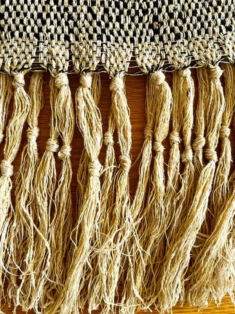 May include: Close-up of a woven textile with a black and beige geometric pattern at the top. Below, long, knotted beige fringe hangs down. The textile is likely a decorative element, such as a throw or wall hanging.