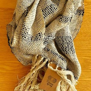 May include: A beige and black woven scarf with a fringed edge. The scarf is folded and draped, showcasing its texture and pattern. A small tag with text is attached to the fringe.