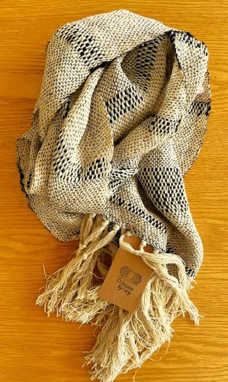 May include: A beige and black woven scarf with a fringed edge. The scarf is folded and draped, showcasing its texture and pattern. A small tag with text is attached to the fringe.