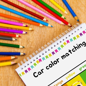 May include: A notebook with the title "Car color matching" and a color chart with the colors orange, yellow, green, and blue. There are colored pencils in a rainbow of colors on a wooden surface.