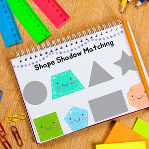 May include: A notebook with a grid pattern and the words "Shape Shadow Matching" written on the cover. The notebook has a variety of colorful shapes with happy faces on them, including a circle, a square, a star, a pentagon, and a trapezoid. There are also some gray shapes that are the shadows of the colorful shapes. The notebook is surrounded by colorful pencils, rulers, and paper clips.