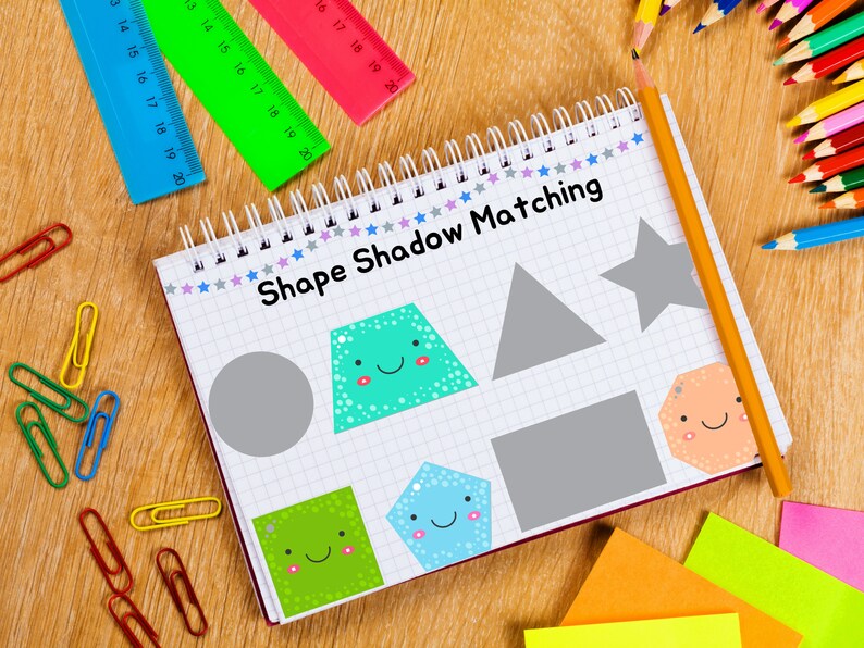 May include: A notebook with a grid pattern and the words "Shape Shadow Matching" written on the cover. The notebook has a variety of colorful shapes with happy faces on them, including a circle, a square, a star, a pentagon, and a trapezoid. There are also some gray shapes that are the shadows of the colorful shapes. The notebook is surrounded by colorful pencils, rulers, and paper clips.