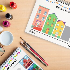 May include: A worksheet with a line of colorful houses of different sizes, labeled "Tallest" and "Shortest".  The worksheet is on a wooden table with paint bottles, paintbrushes, and a bowl.  The text "Sorting by size" is at the top of the worksheet.