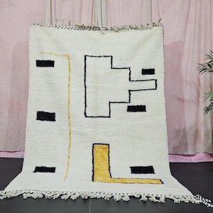 Könnte beinhalten: Cremefarbener Teppich mit geometrischem Design. Der Teppich hat schwarze und gelbe Formen auf weißem Hintergrund. Er hat einen Fransenbesatz am unteren Rand und wird vor einem rosa Vorhang präsentiert. Ungefähre Größe: 150 cm x 200 cm.