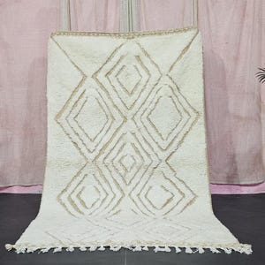 Könnte beinhalten: Cremefarbener, rechteckiger Teppich mit einem geometrischen Rautenmuster in hellem Beige. Der Teppich hat einen dicken, strukturierten Flor und einen Fransenbesatz am unteren Rand. Der Teppich wird vor einem rosa Vorhang präsentiert.