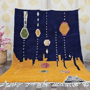 May include: A hand-woven rug with a navy blue background and a mustard yellow base. The rug features geometric shapes in white, green, yellow, and pink. The rug has a fringe along the bottom edge.