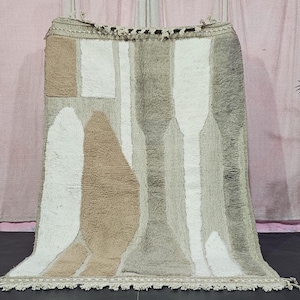 Könnte beinhalten: Ein rechteckiger Teppich mit geometrischem Design in Beige-, Weiß- und Grautönen. Der Teppich hat einen Fransenbesatz am unteren Rand und eine strukturierte Oberfläche. Das Design umfasst abstrakte Formen und vertikale Streifen.