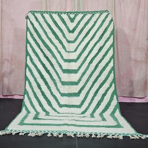 Könnte beinhalten: Ein rechteckiger Teppich mit einem geometrischen Muster aus grünen und weißen Chevron-Streifen. Der Teppich hat einen Fransenbesatz am unteren Rand. Der Teppich wird vor einem rosa Vorhanghintergrund präsentiert.