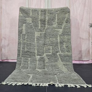 Könnte beinhalten: Ein grauer, strukturierter Teppich mit geometrischem Muster. Der Teppich hat eine rechteckige Form mit Fransen am unteren Rand. Das Design zeigt erhabene, rechteckige Formen in verschiedenen Größen. Der Teppich wird vor einem rosa Vorhang präsentiert.