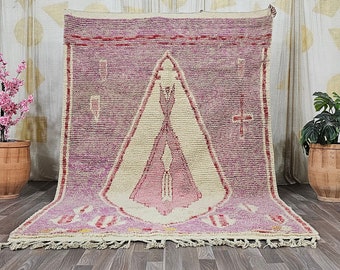 Puede incluir: Una alfombra marroquí rosa y crema con un patrón geométrico. La alfombra tiene un borde de flecos y se muestra en un suelo de madera.