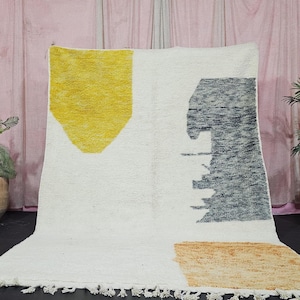 Könnte beinhalten: Ein weißer Teppich mit einem geometrischen Design mit gelben, grauen und beigen Formen. Der Teppich hat einen Fransenrand an der Unterkante und wird vor einer rosa Wand mit Topfpflanzen auf beiden Seiten präsentiert.