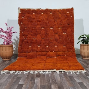Puede incluir: Una alfombra marroquí grande, de color naranja, tejida a mano con un patrón geométrico. La alfombra tiene una franja blanca alrededor de los bordes y está colocada sobre un suelo de madera.