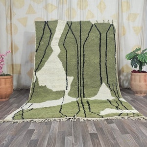 Puede incluir: Una alfombra de estilo abstracto con patrones verdes y blancos y líneas negras. La alfombra tiene un borde de flecos y está colocada sobre un suelo de madera.