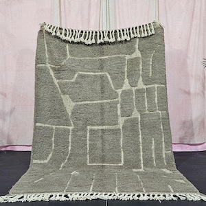 Könnte beinhalten: Ein großer, olivgrüner Teppich mit einem geometrischen Muster aus cremefarbenen Linien. Der Teppich hat eine strukturierte Oberfläche und einen Fransenbesatz an den oberen und unteren Kanten. Der Teppich wird vor einem rosa Vorhang präsentiert.