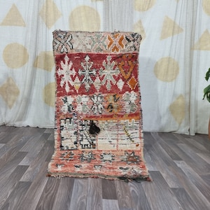 May include: A colorful Moroccan rug with geometric patterns in shades of red, orange, white, and black. The rug is folded and standing upright on a wooden floor.
