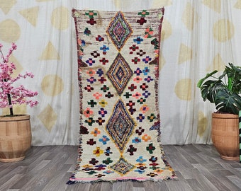 May include: A rectangular, cream-colored rug with a central diamond pattern in shades of blue, red, and yellow. The rug features a repeating pattern of colorful floral and geometric shapes. The rug is displayed on a wooden floor.