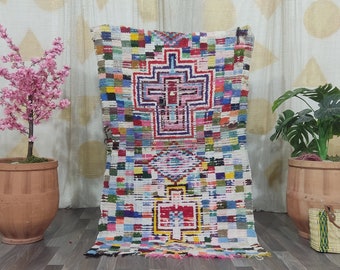 May include: A colorful Moroccan rug with a geometric pattern in shades of pink, blue, green, yellow, and red. The rug is laid out on a wooden floor in front of a white wall with a geometric pattern.