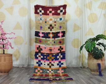 May include: A colorful Moroccan rug with geometric patterns in shades of pink, blue, green, red, and white. The rug is laid out on a wooden floor in a room with white walls and a potted plant.