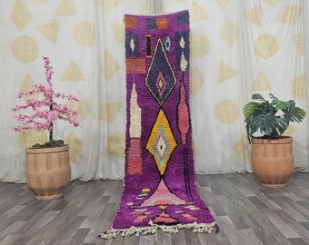 May include: A vibrant, rectangular, purple rug with geometric patterns in yellow, pink, and blue. The rug has a diamond and rectangular design. The rug is displayed on a wooden floor with a neutral background.
