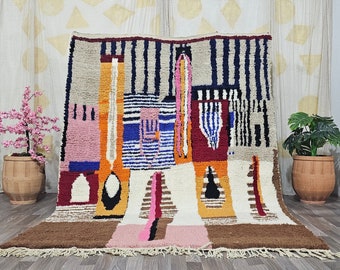May include: A colorful, abstract Moroccan rug with geometric patterns in shades of orange, blue, pink, and brown. The rug has a fringe border and is woven with a variety of textures.