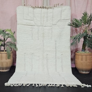 Könnte beinhalten: Cremefarbener, rechteckiger Teppich mit geometrischem Muster. Der Teppich hat eine strukturierte Oberfläche und einen Fransenbesatz am unteren Rand. Zwei Topfpflanzen flankieren den Teppich und verleihen ihm einen Hauch von Grün. Der Teppich wird vor einem rosa Hintergrund präsentiert.