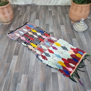 May include: A rectangular woven rug featuring a repeating diamond pattern in vibrant colors including red, yellow, blue, pink, and black against a cream background. The rug has a textured pile and dark fringe detailing at one end.