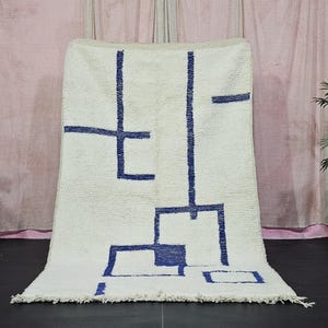 Könnte beinhalten: Cremefarbener Teppich mit einem geometrischen Design in Marineblau. Der Teppich zeigt eine Reihe von verbundenen Quadraten und Linien, die ein abstraktes Muster bilden. Er wird vor einem neutralen Hintergrund präsentiert, der seine strukturierte Oberfläche und sein Design hervorhebt.