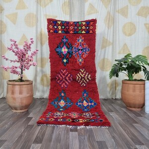 May include: A rectangular runner rug featuring a deep red base color with a high-pile texture. The rug is decorated with symmetrical geometric patterns in vibrant shades of blue, yellow, pink, and green, arranged in horizontal rows across the surface.