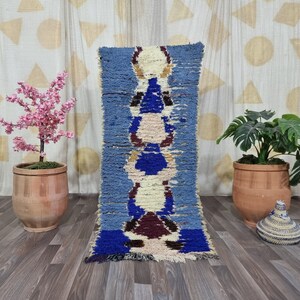 May include: A rectangular, blue and white Moroccan rug with a central pattern of stacked shapes in cream, blue, and burgundy. The rug is displayed on a wooden floor, with potted plants and a patterned curtain in the background.