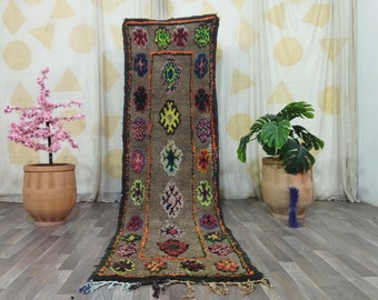 May include: A long, rectangular, brown rug with a repeating pattern of colorful floral and geometric designs. The rug has a dark border with orange and green accents. The rug is displayed on a wooden floor.