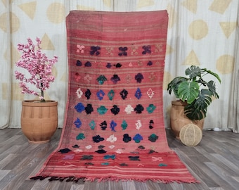 May include: A red and pink vintage rug with a geometric pattern of black, blue, and green shapes. The rug has a fringe border and is laid out on a wooden floor.