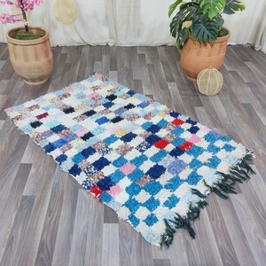 Puede incluir: Una alfombra marroquí colorida con un patrón a cuadros en tonos de azul, blanco, rosa, rojo y marrón. La alfombra tiene un borde de flecos y está colocada sobre un suelo de madera.