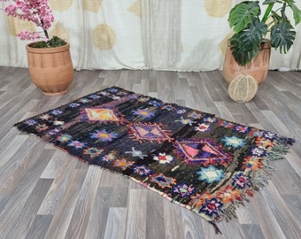 May include: A colourful Moroccan rug with a black background and intricate diamond patterns in shades of pink, blue, yellow, and orange. The rug has a fringed border and is laid out on a wooden floor.