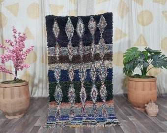 May include: A rectangular, hand-woven rug with a geometric diamond pattern. The rug features a color palette of black, brown, blue, green, and gray. The rug is displayed indoors, with a wooden floor and decorative plants in the background.