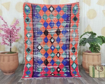 May include: A vibrant, rectangular rug with a geometric diamond pattern in shades of orange, blue, green, and black. The rug has a fringed edge and is displayed indoors, suggesting a decorative home accent.
