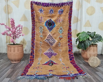 May include: A Moroccan rug with a brown background and a diamond pattern in shades of blue, pink, and purple. The rug has a fringe border in red, purple, and blue.