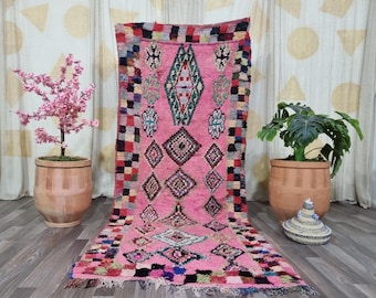 May include: A pink, rectangular rug with geometric patterns. The rug features diamond and square designs in various colors, including pink, green, blue, and red. The rug is displayed on a wooden floor.